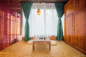 Una habitación con suelo de madera, una mesa con lámpara colgante, cortinas verdes y un armario de madera.