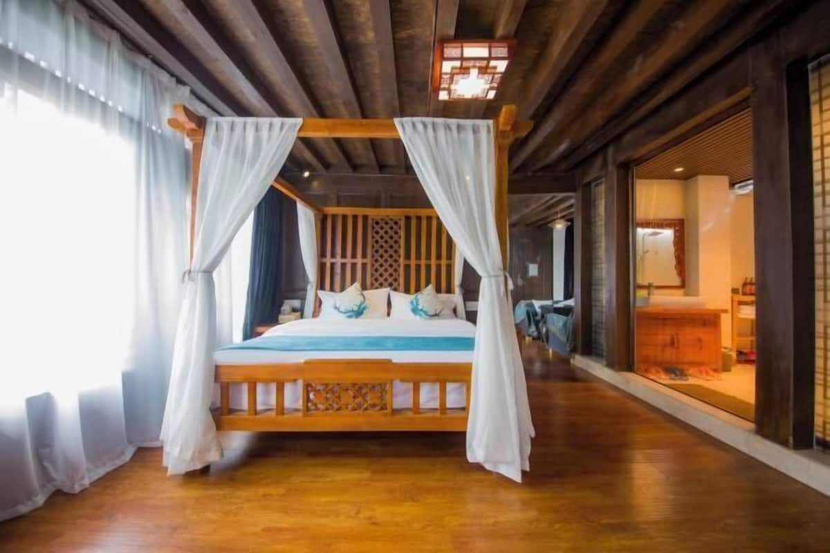 A wooden four-poster bed with white drapes, a wooden floor, and a traditional wooden partition in the background.