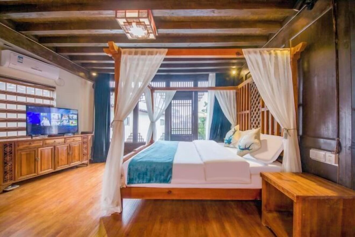 Quarto com cama de madeira, cortinas brancas, uma TV em um armário de madeira e vista para o exterior.