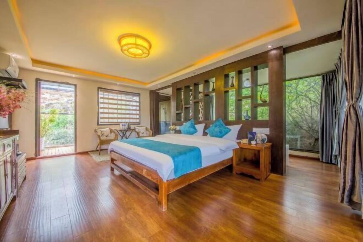 A spacious bedroom with a large bed, wooden flooring, a built-in bookshelf, and large windows.