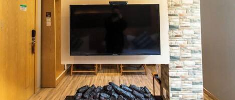A hotel room with a large flat-screen TV mounted on a stone wall, a wooden door, and a fireplace with logs.