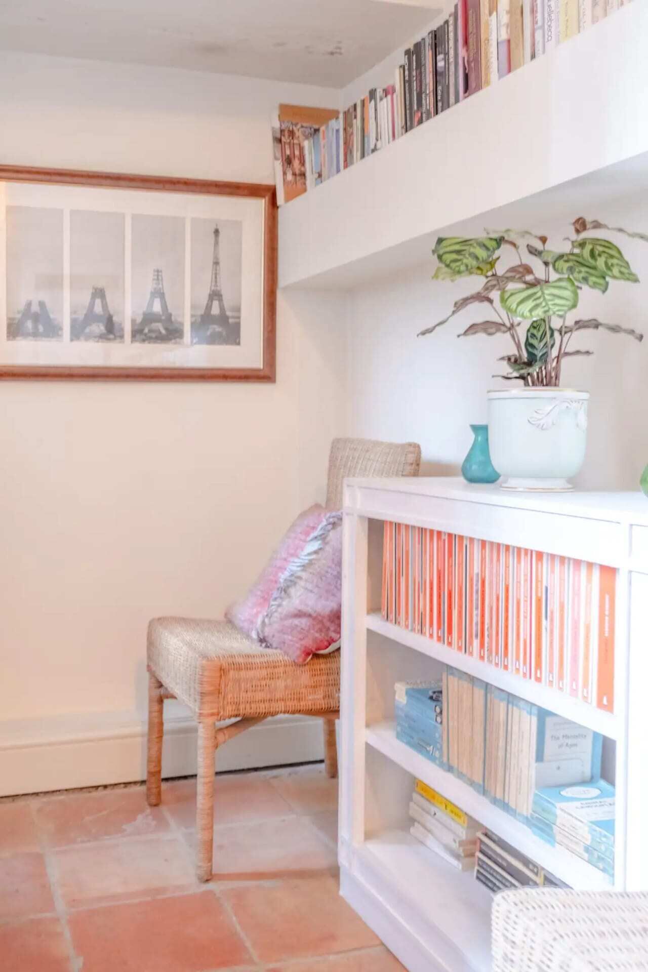 A cozy reading nook with a wicker chair, a bookshelf filled with books, a framed picture of the Eiffel Tower, and a potted plant.