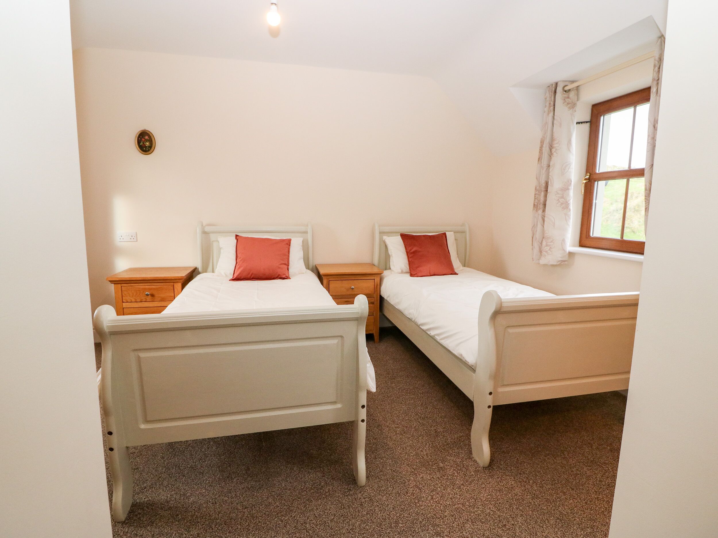 A bedroom with two single beds, each with a nightstand and a lamp. There is a window with curtains and a wall-mounted light fixture.
