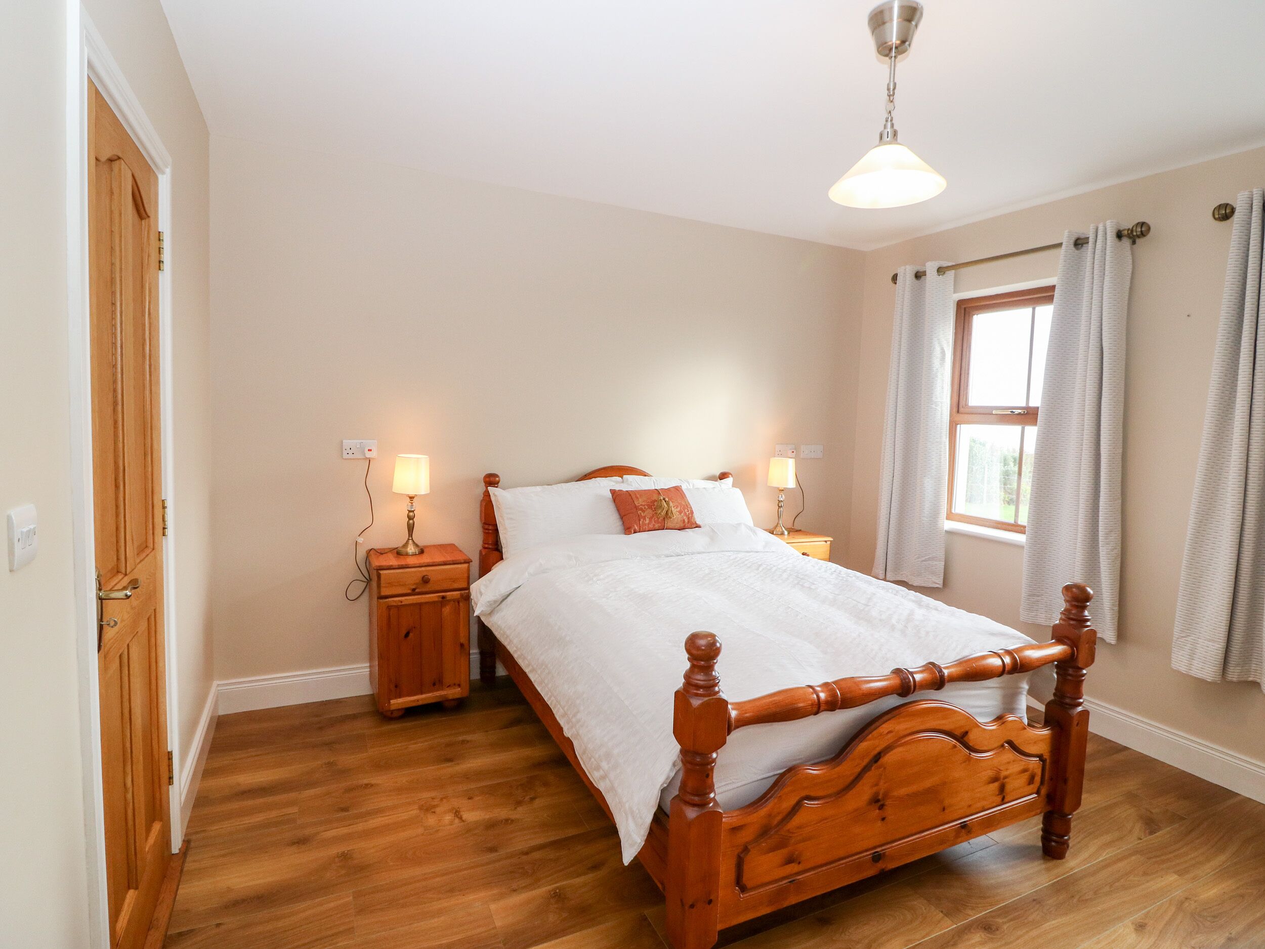 A bedroom with a wooden bed, two bedside tables with lamps, a window with curtains, and a wooden floor.