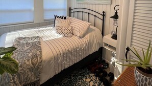 Une chambre à coucher comprenant un lit, un ventilateur de plafond, une fenêtre avec des stores, une lampe fixée au mur et une plante en pot.