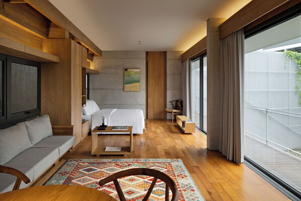 A modern hotel room with a large bed, a sofa, a wooden coffee table, and a view of the outdoors through a large window.