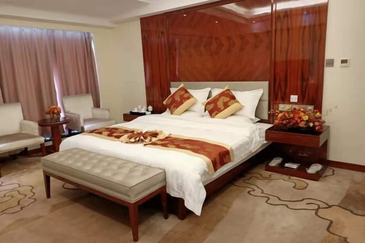 A hotel room with a large bed, a wooden headboard, a bedside table with a fruit basket, a chair, and a small table with a vase.