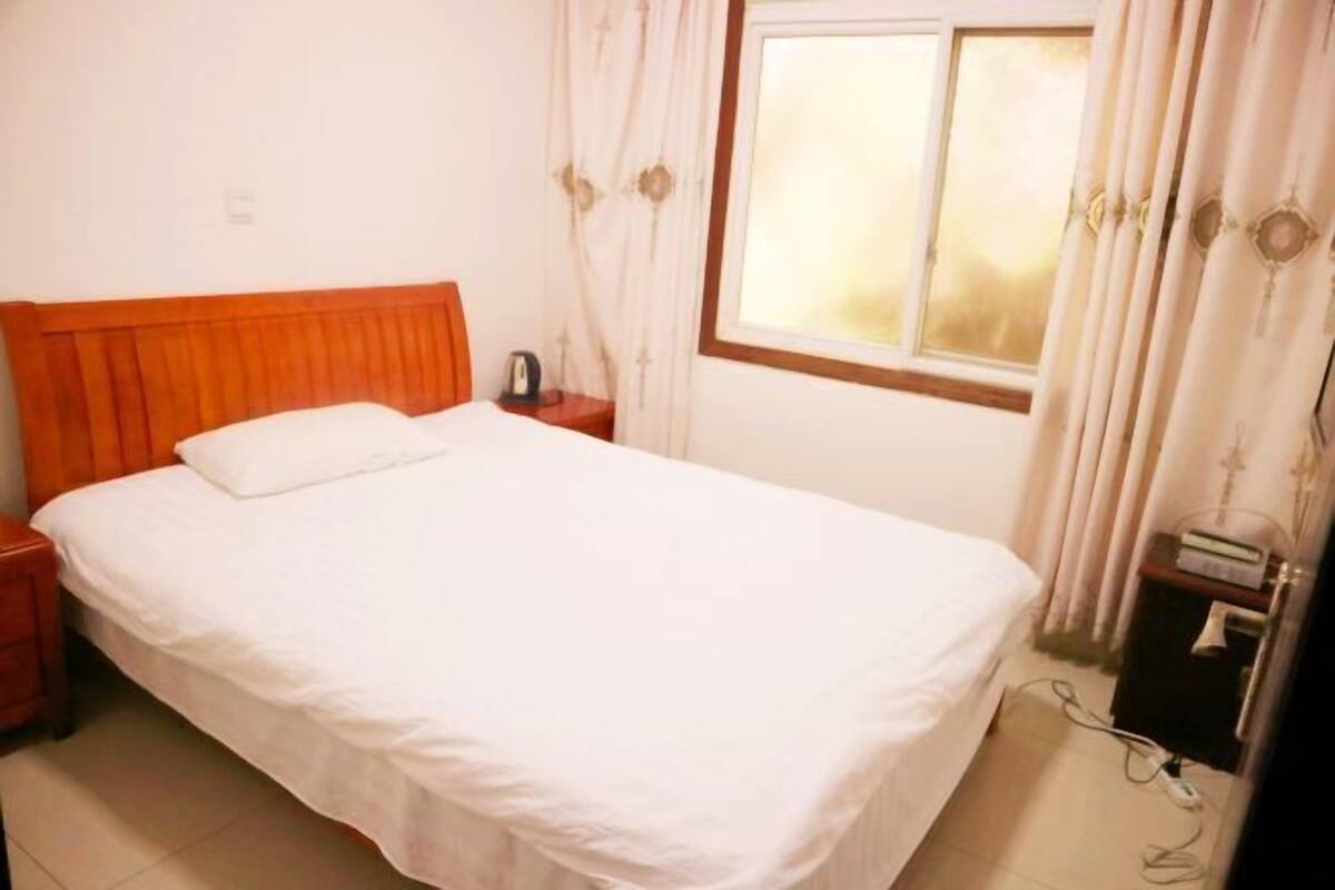 A single bed with a wooden headboard, a bedside table, a nightstand, a window with curtains, and a wall socket.