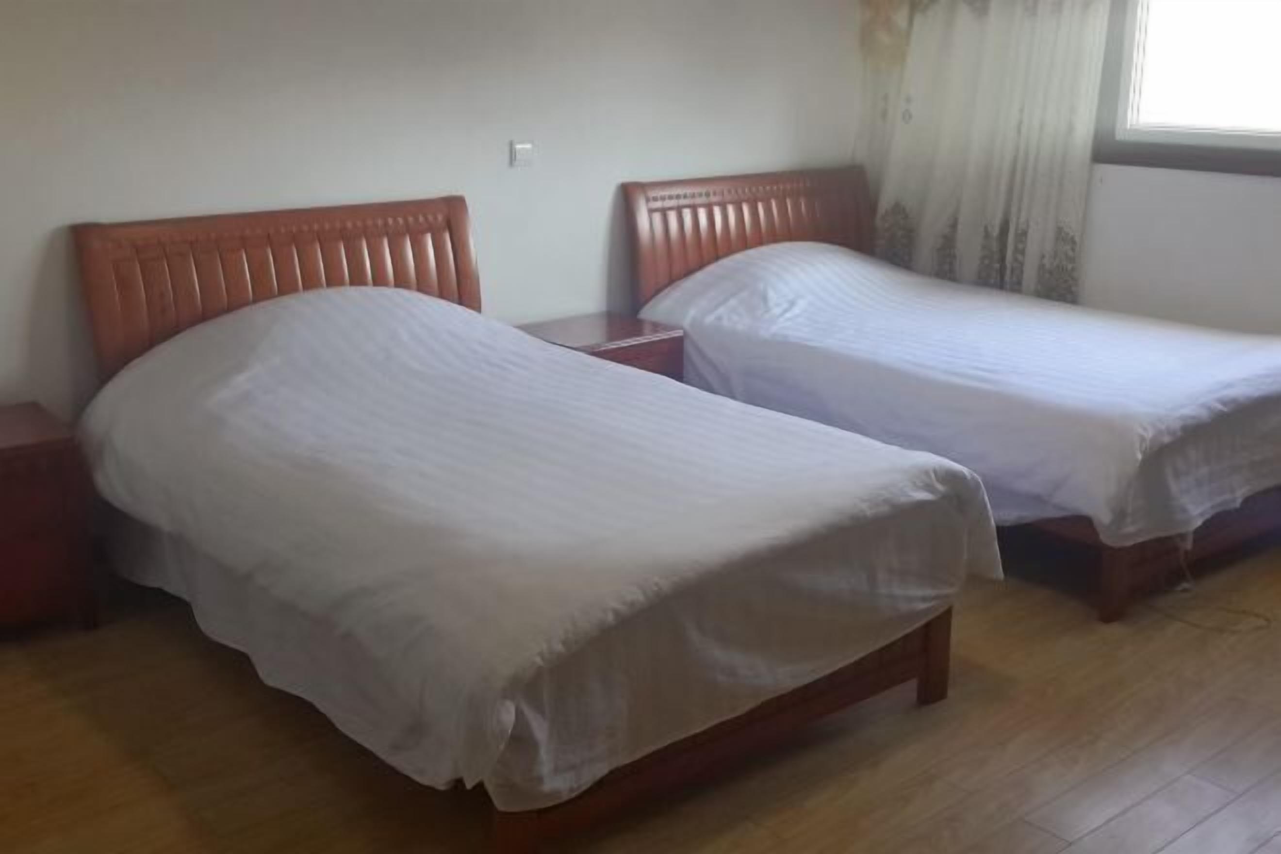 A room with two single beds, wooden headboards, a bedside table, and a window with curtains.
