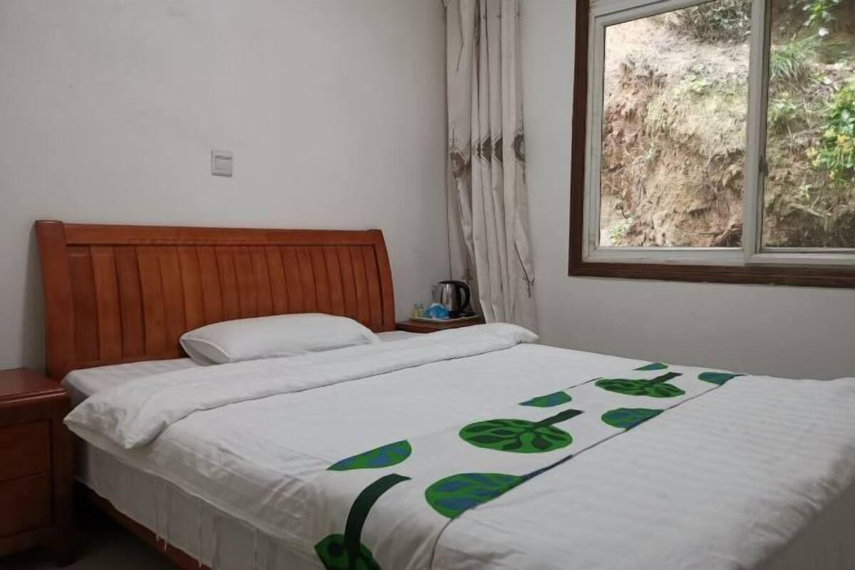 A single bed with a wooden headboard, a white bedspread with green leaf patterns, a nightstand, a window with a view of a rocky outdoor area, and a curtain partially drawn.