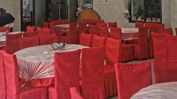 Un salón de banquetes con mesas redondas y sillas rojas, una silla de madera y un ventanal con cortinas.