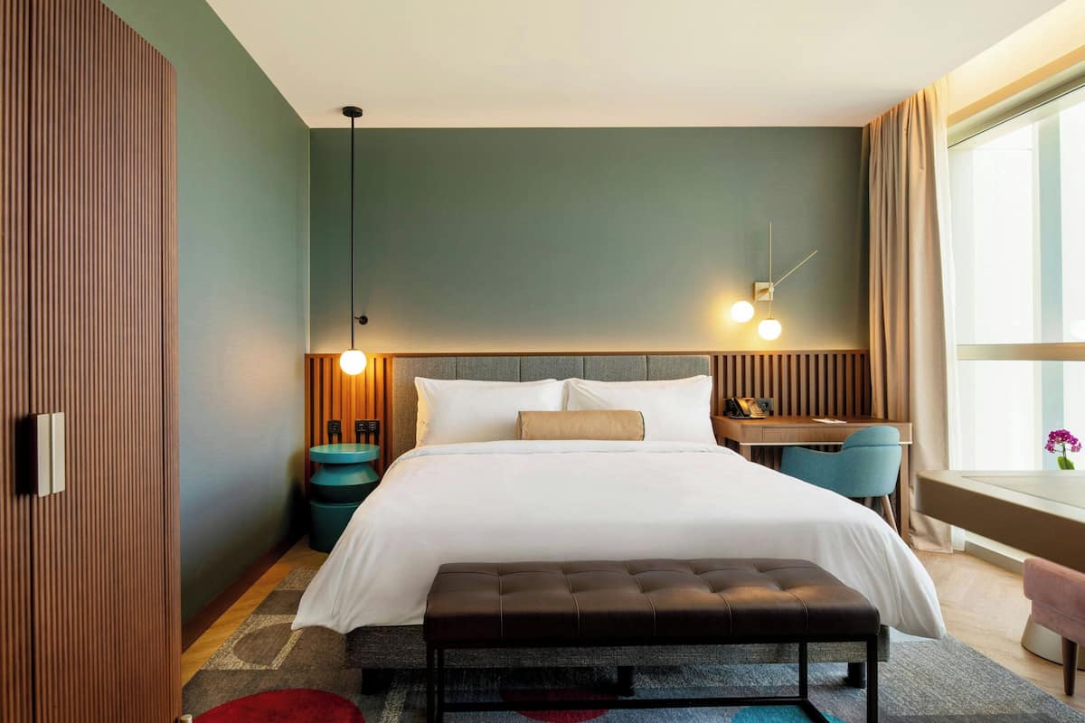 A modern hotel room with a large bed, a desk, a bench, and a view of the city.
