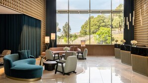Un vestíbulo moderno con un sofá verde azulado curvado, una mesa de centro redonda y un área de estar con sillones. Ventanas amplias ofrecen vistas al exterior con vegetación.