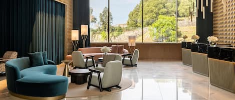 A modern lobby with a curved teal sofa, a round coffee table, and a seating area with armchairs. Large windows offer a view of greenery outside.