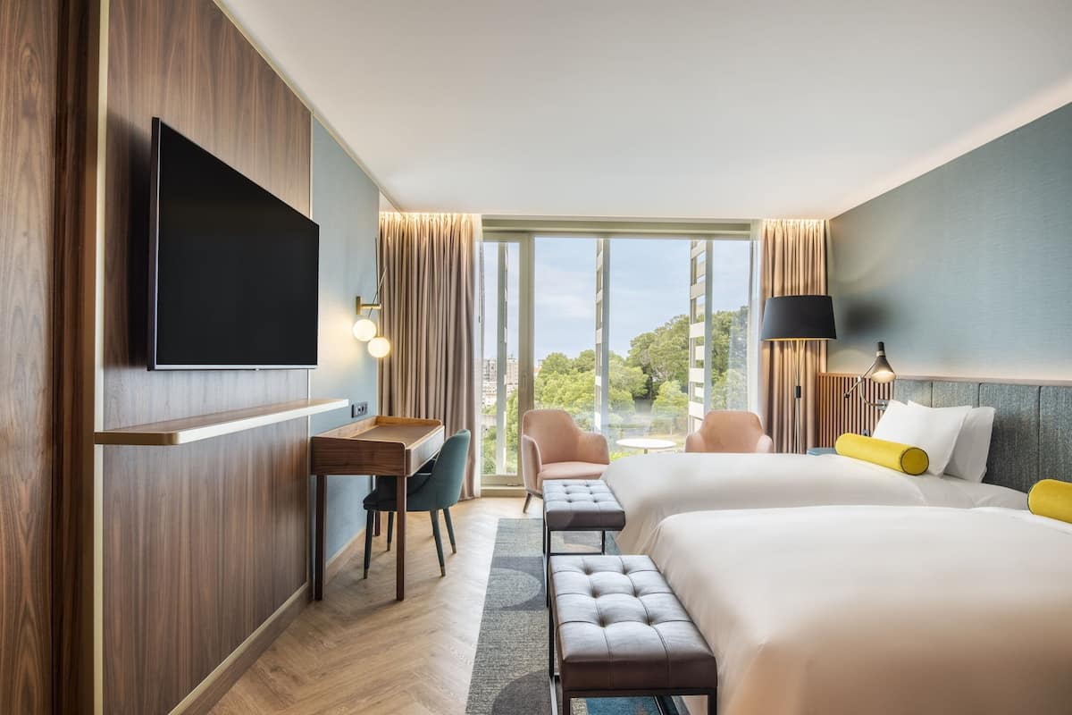 A hotel room with a large TV, a desk, two beds, a bench, and a view of the outdoors.