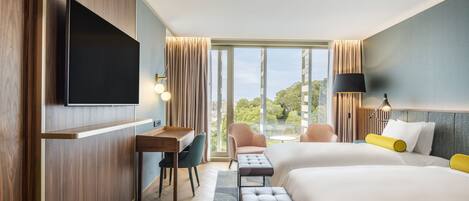 Habitación de hotel con televisor grande, escritorio, dos camas, un banco y vistas al exterior.