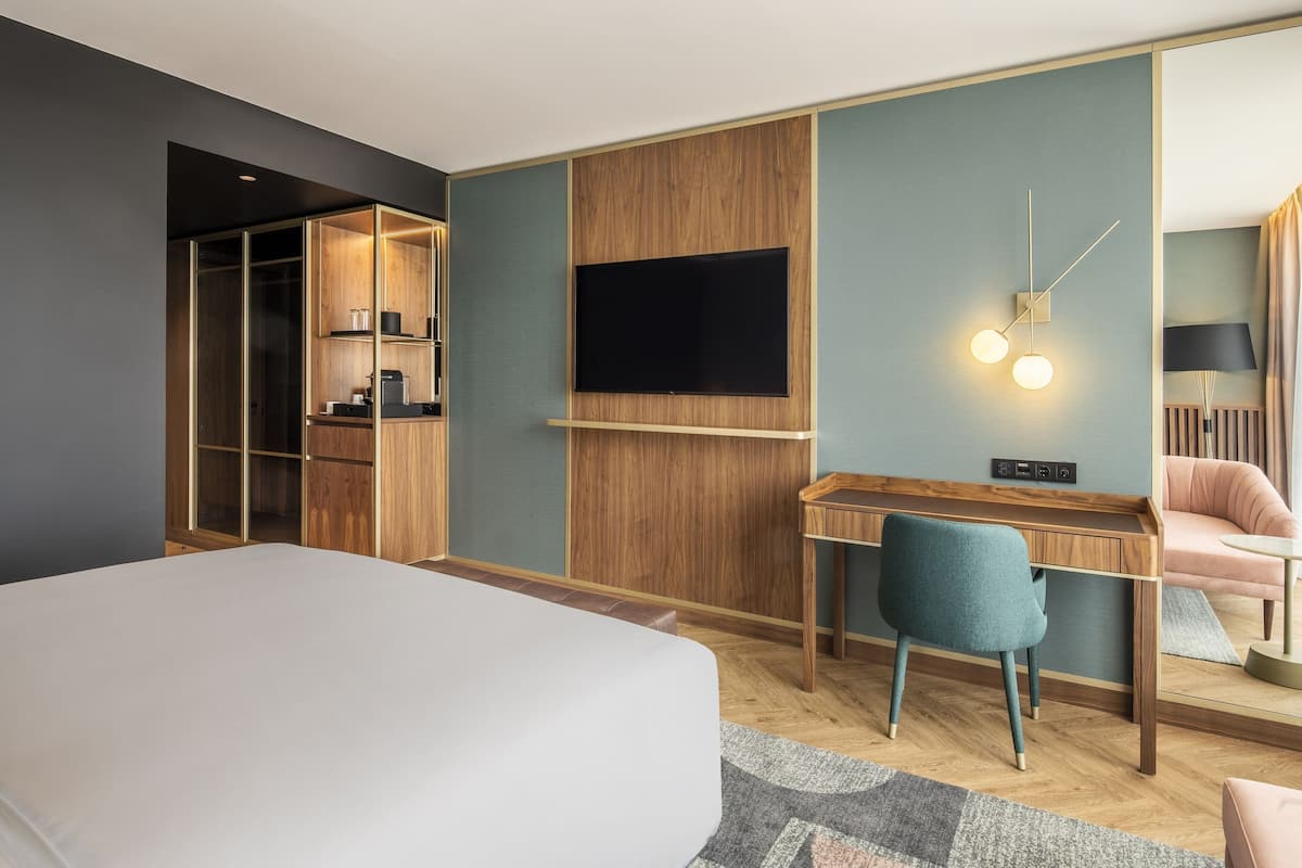 A modern hotel room with a large bed, a desk with a chair, a TV mounted on the wall, and a wardrobe with a mirror.