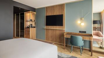 A modern hotel room with a large bed, a desk with a chair, a TV mounted on the wall, and a wardrobe with a mirror.