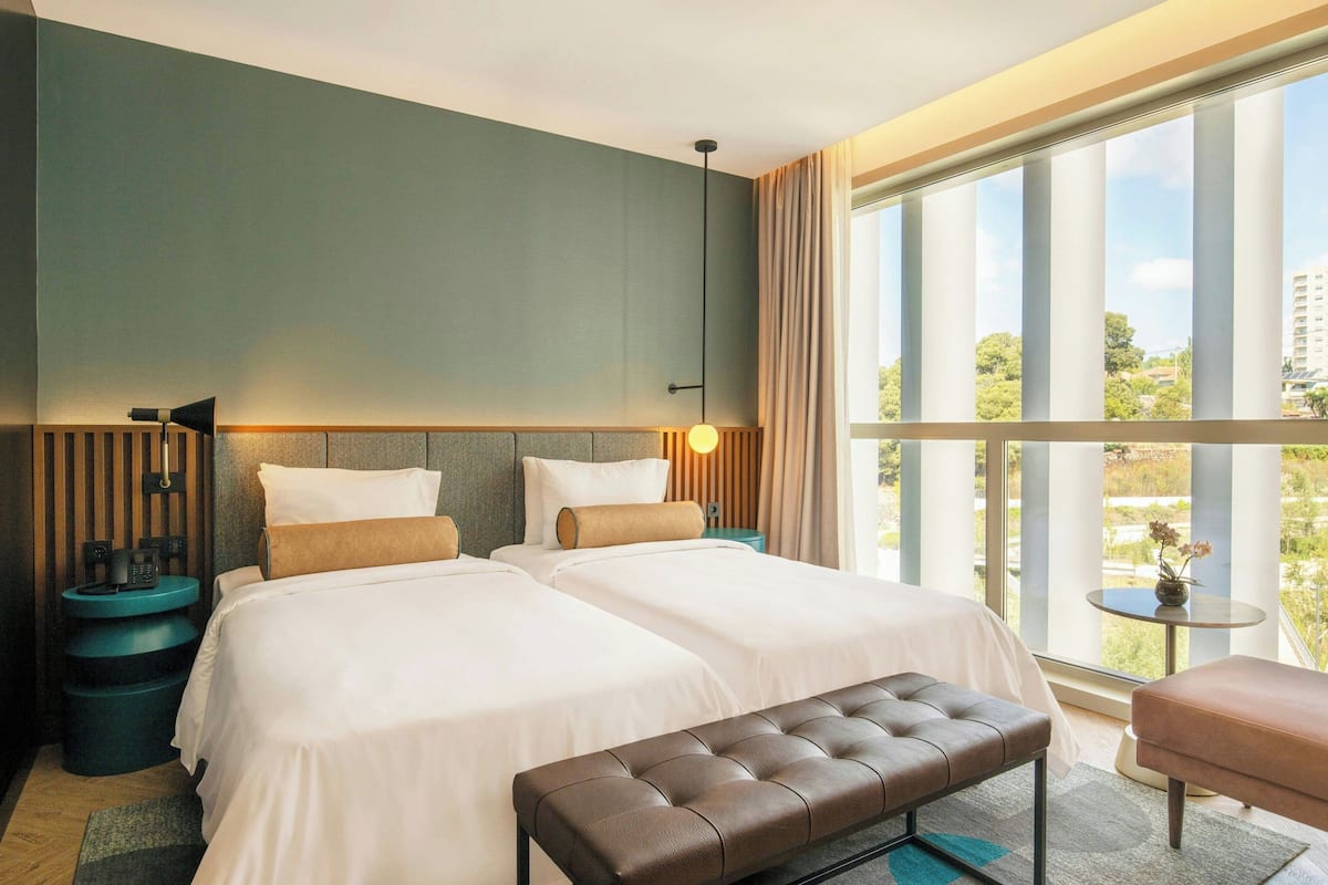 A hotel room with two beds, a bench, a small table, and a view of greenery through large windows.