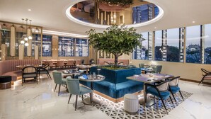 Un interior moderno con una zona de estar central, un árbol grande y vistas de la ciudad a través de amplias ventanas.