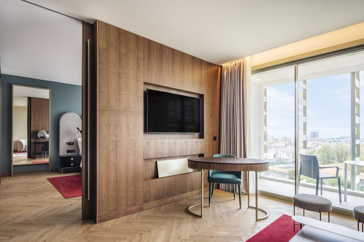 A modern hotel room with a large TV mounted on a wooden wall, a round dining table with chairs, and a large window offering a city view.