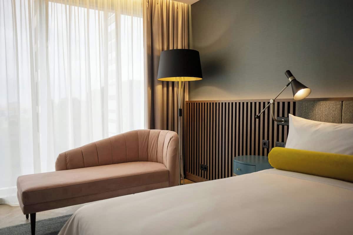 A hotel room with a bed, a chair, a bedside table, a lamp, and a window with curtains.