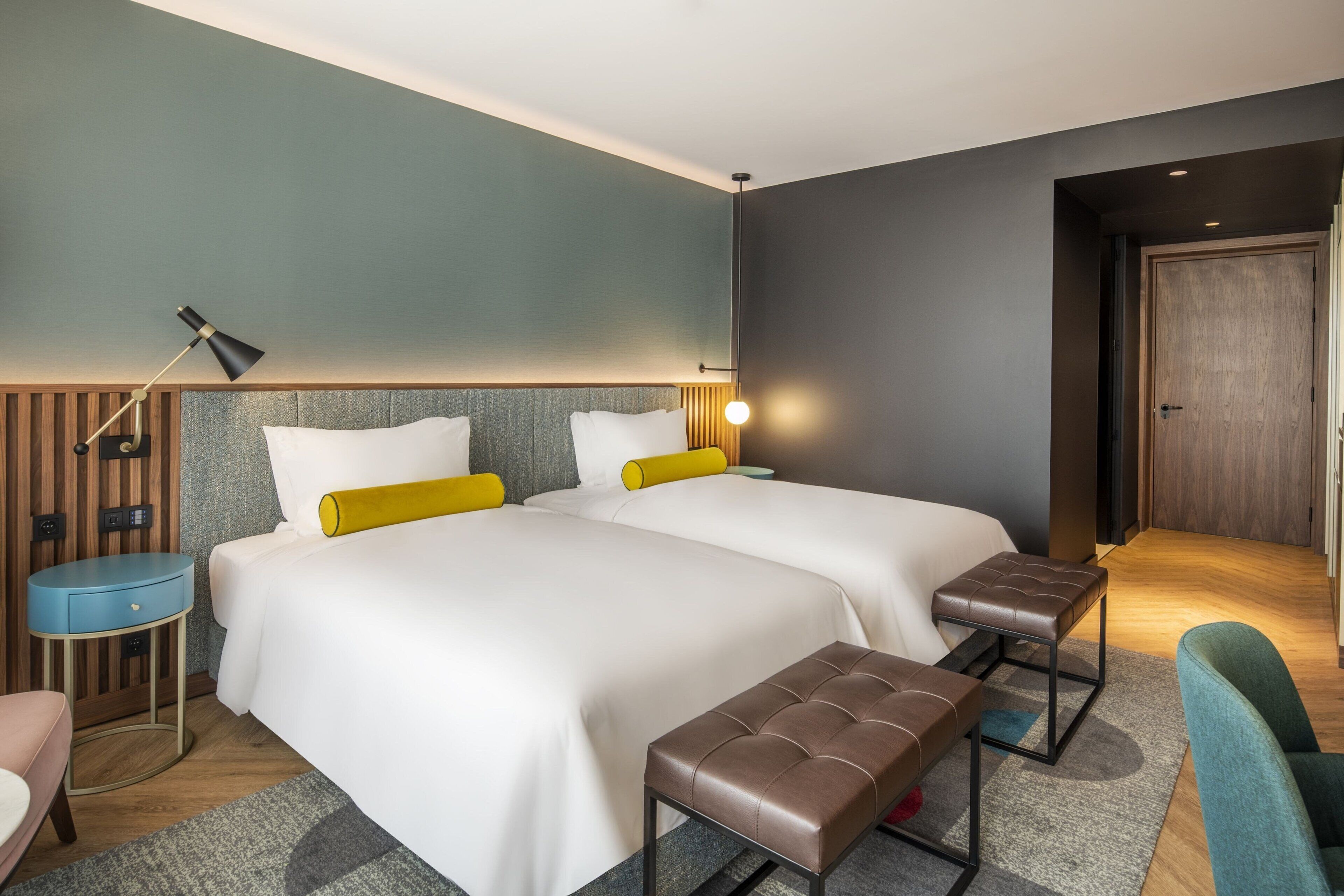 A hotel room with two beds, a desk, a chair, a bench, a lamp, and a door.