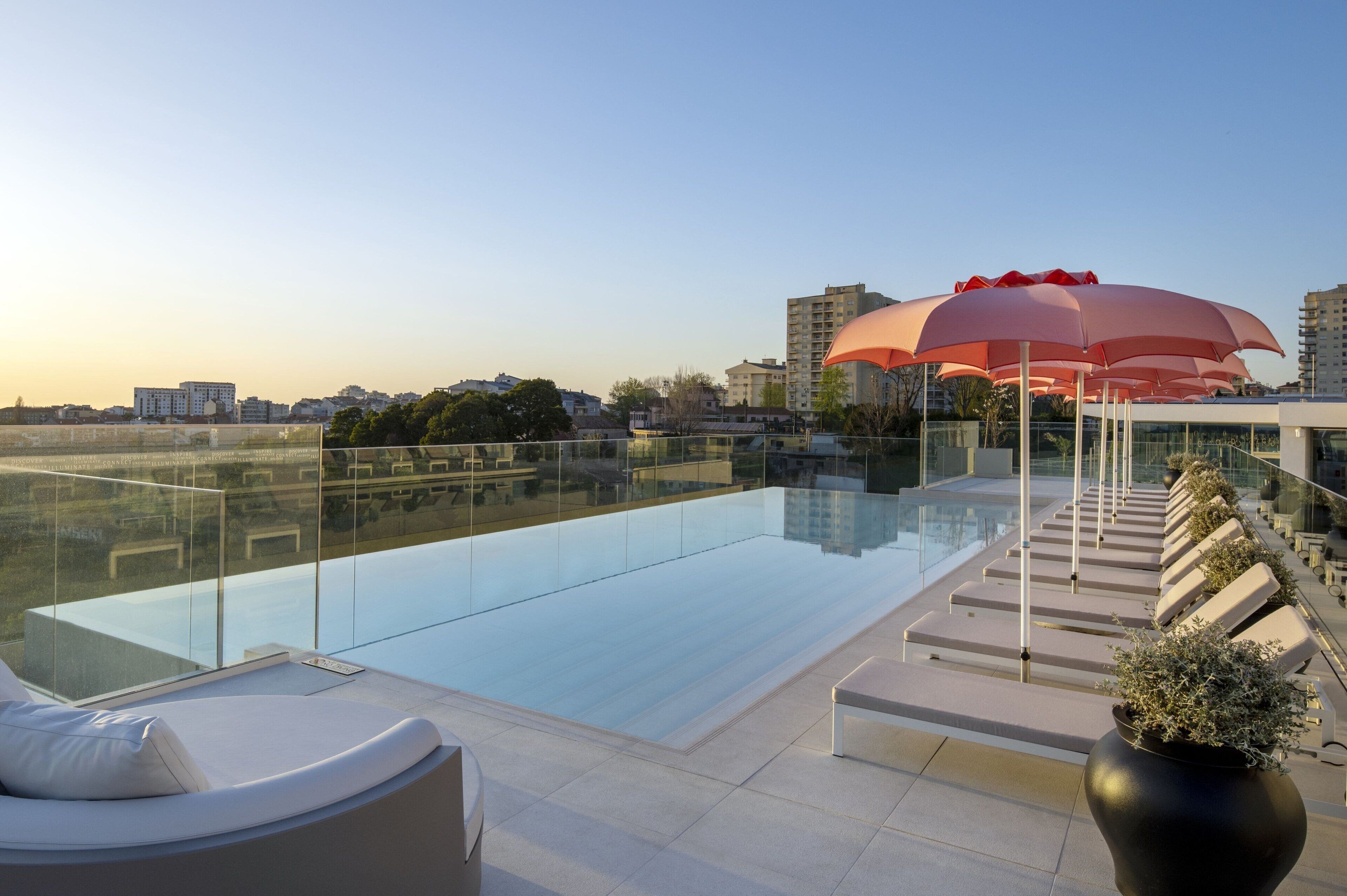 A rooftop infinity pool with glass railings, lounge chairs, and a row of red umbrellas.