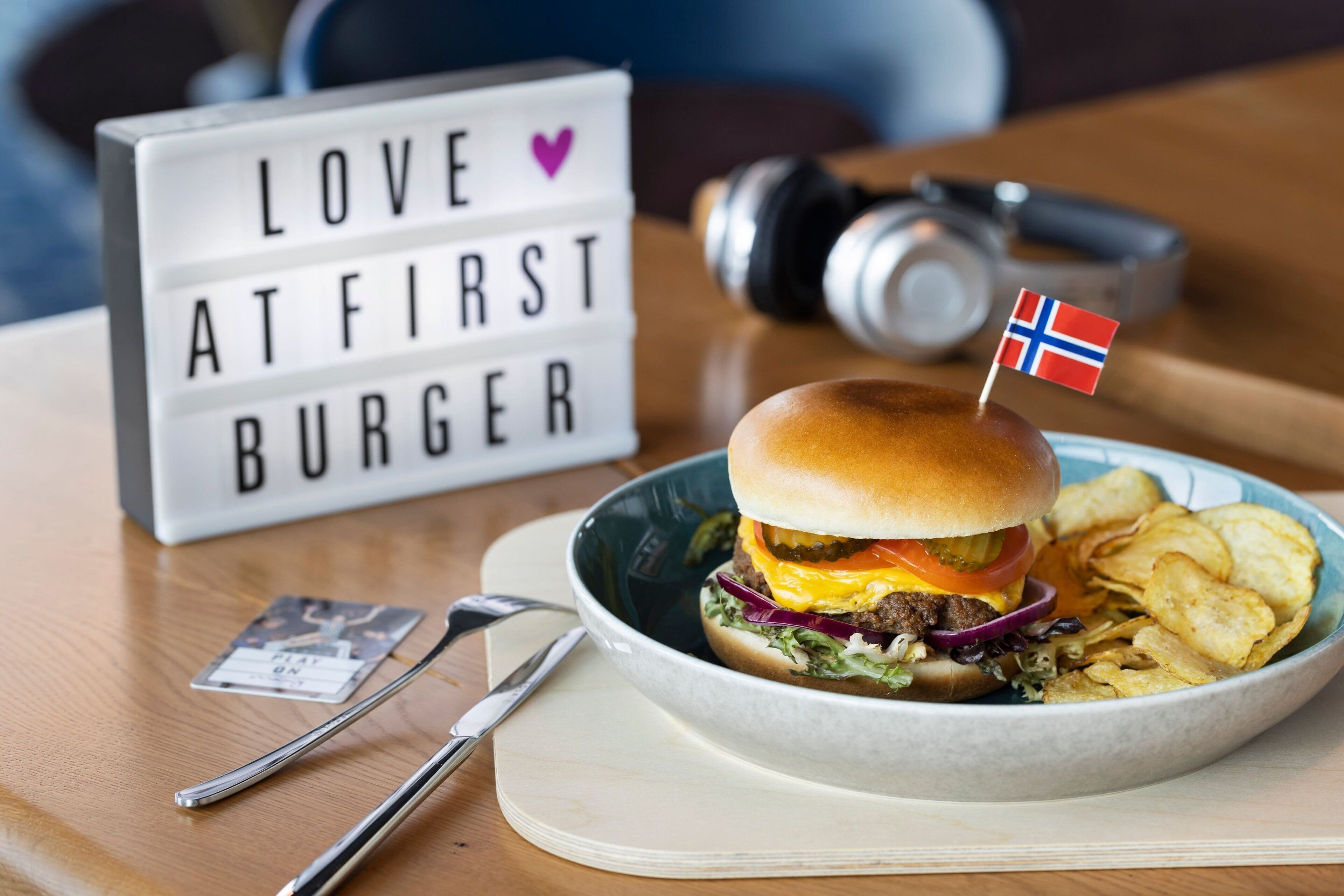 Un hamburger orné d’un drapeau, une assiette de frites, une boîte lumineuse avec un message, et une paire d’écouteurs posés sur une table.