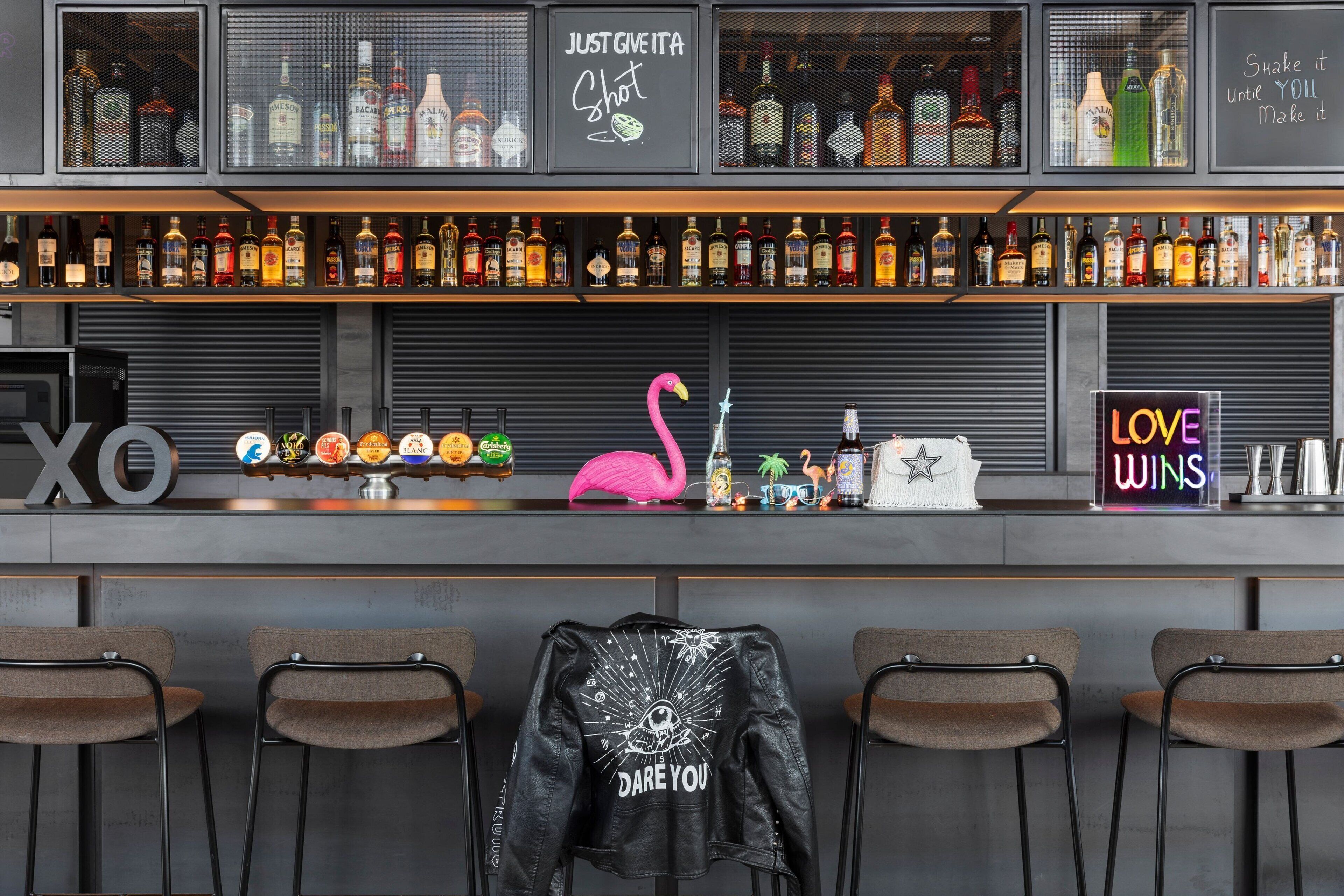 Un bar avec des étagères remplies de bouteilles d'alcool de toutes sortes, un panneau au néon représentant un flamant rose et un autre panneau au néon sur lequel on peut lire : « L’AMOUR TRIOMPHE ».