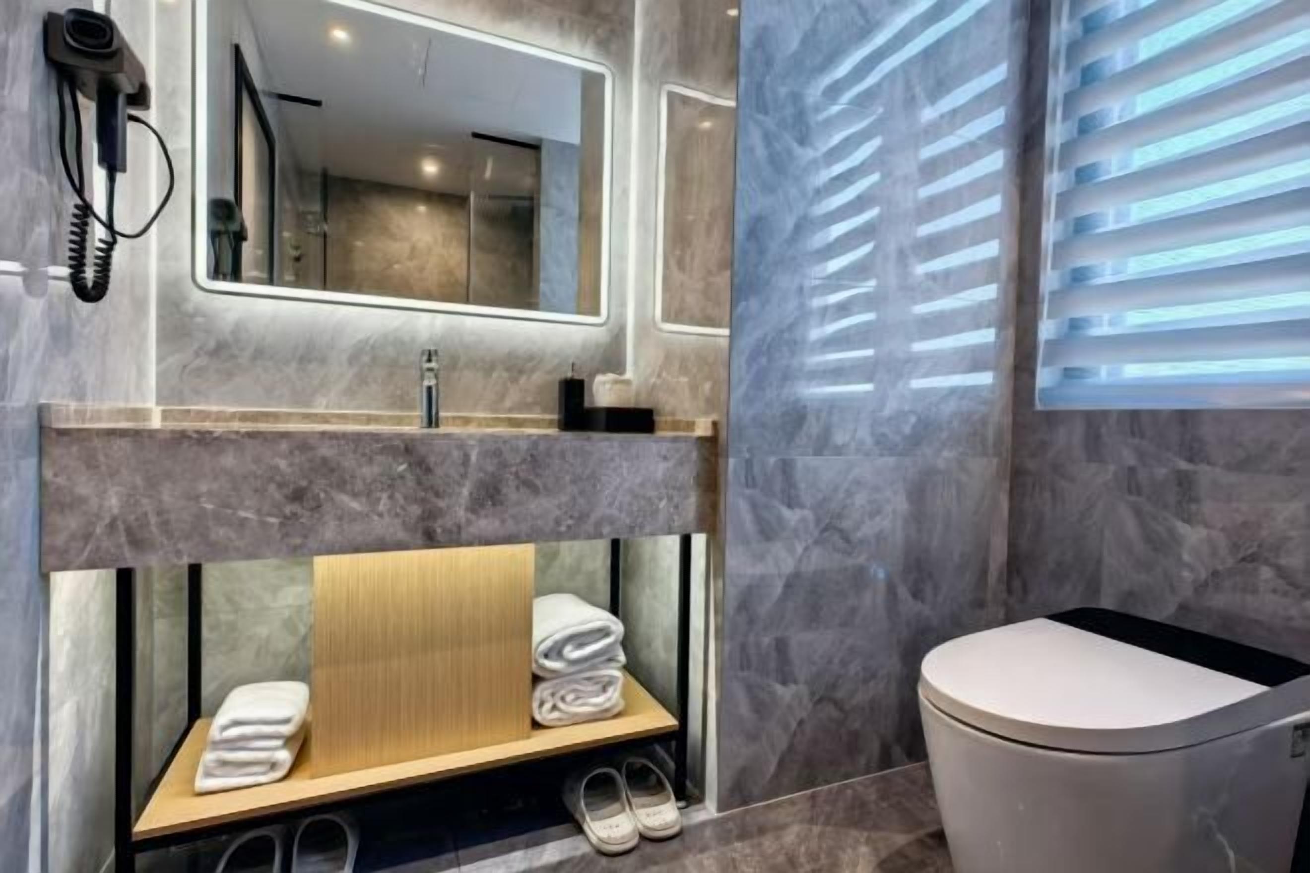 A modern bathroom with a marble vanity, a mirror with integrated lighting, a toilet, and a window with blinds.