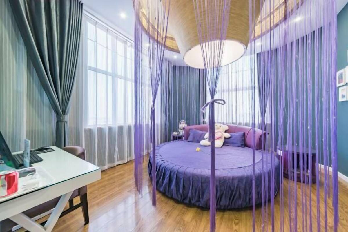 A bedroom with a round bed, a desk, and a large window.