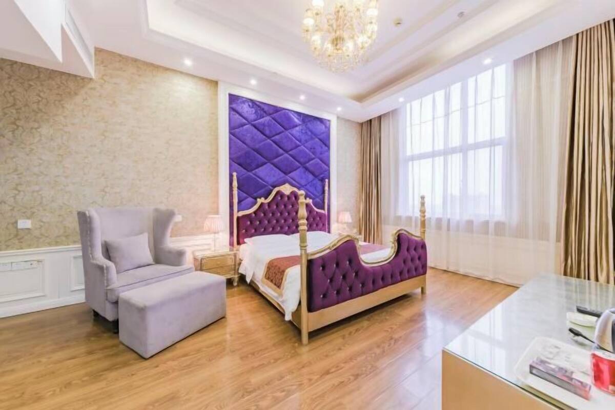 A bedroom with a large bed, a chaise lounge, a glass table, and a chandelier.