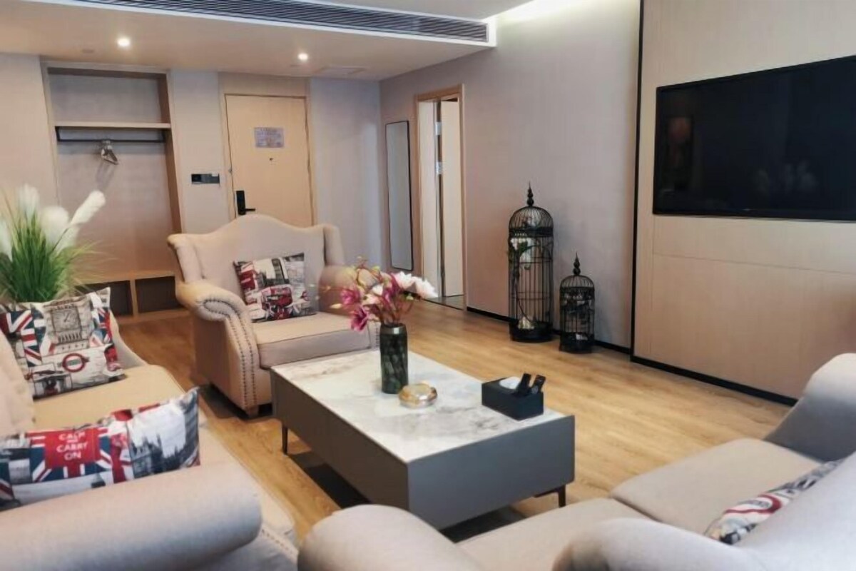 A modern living room with a sofa, armchair, and coffee table. There is a television mounted on the wall and a door in the background.
