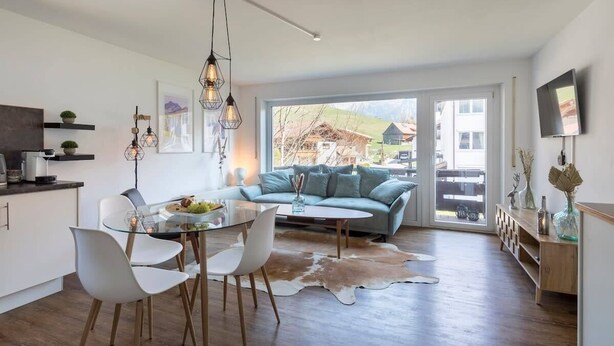 A modern living room with a glass dining table, white chairs, a light blue sofa, and a cowhide rug. - Apartment Alpenglühen nahe Breitachklamm (Oberstdorf)