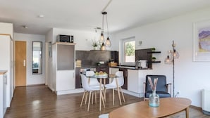 A modern kitchen with white cabinets, a dining area with a glass table and white chairs, and a living area with a dark armchair and a floor lamp. - Apartment Alpenglühen nahe Breitachklamm (Oberstdorf)