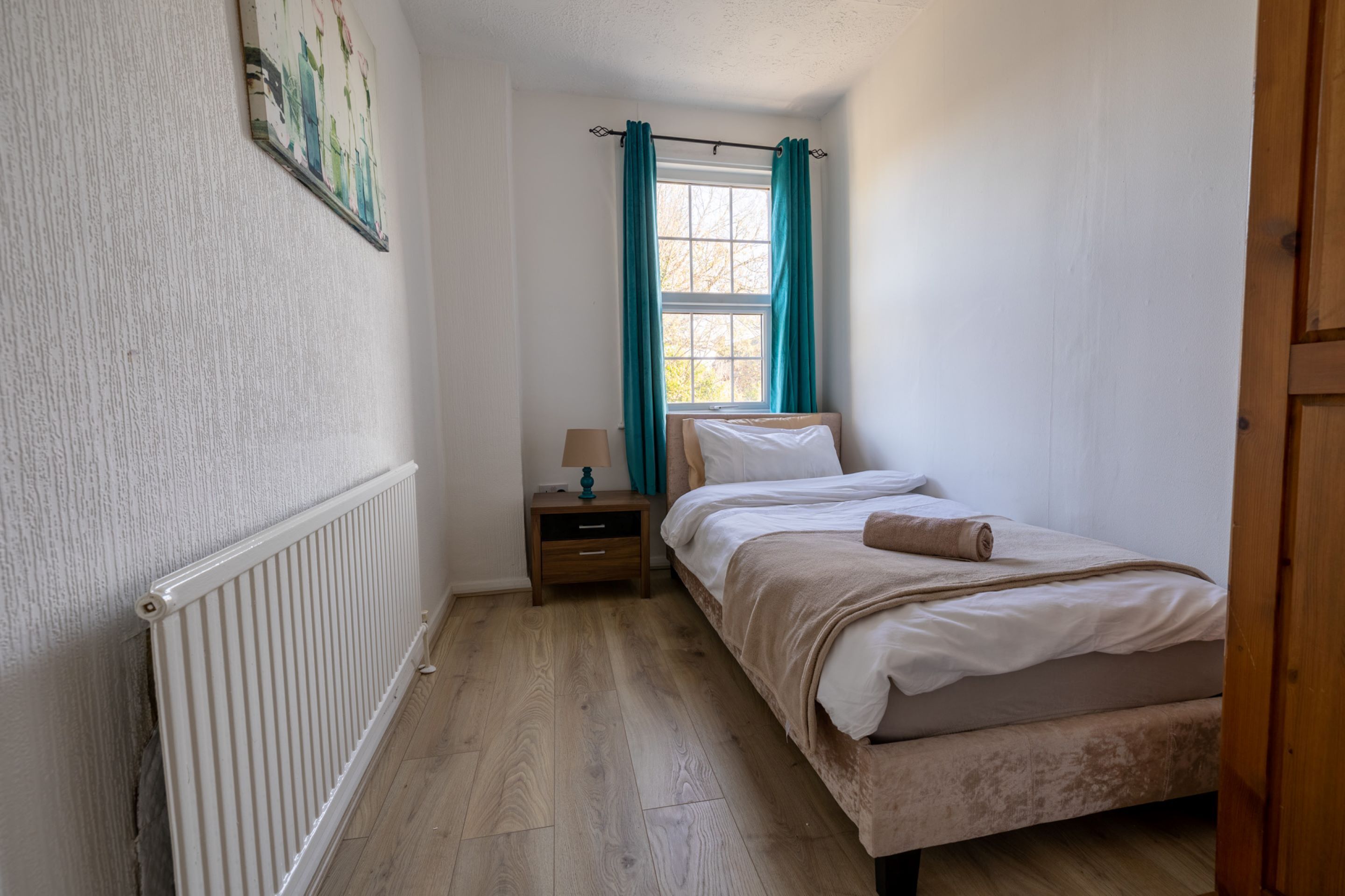 A bedroom with a wooden floor, a bed with white linens, a nightstand with a lamp, and a window with teal curtains.