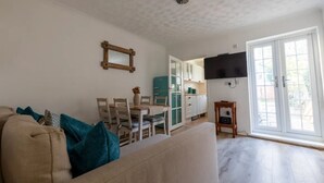 A living room with a sofa, a dining table with chairs, a kitchen area with cabinets, and a TV mounted on the wall. - West Dartford Teal House (Dartford)