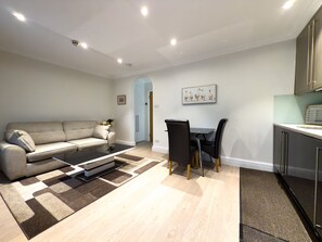 A modern living room with a sofa, a dining table with chairs, a kitchen area, and a hallway. - Chic 1 bedroom apartment in Marylebone (London)