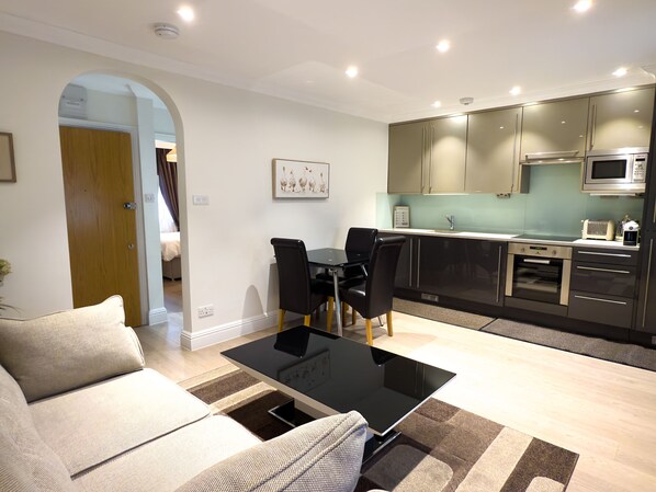 Una sala moderna con una mesa de centro de vidrio negro, un sofá color beige y un comedor con sillas negras. La cocina cuenta con electrodomésticos de acero inoxidable y un salpicadero de color verde claro.