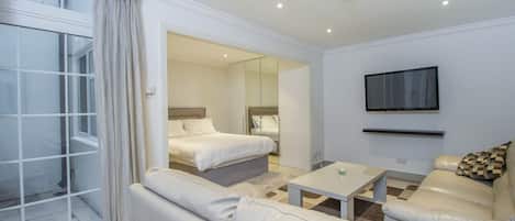A modern hotel room with a large window, a sofa, a bed, a TV, and a mirror.