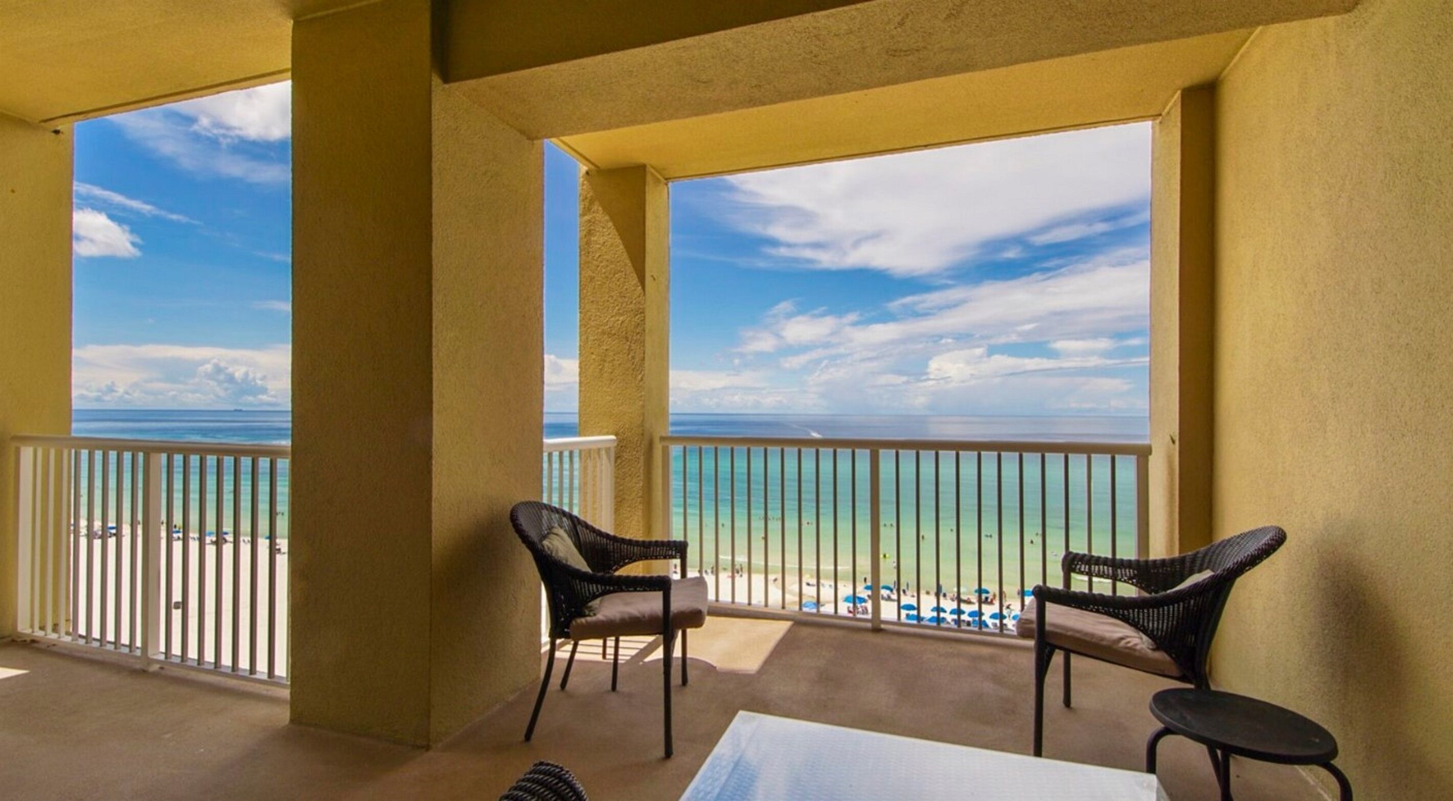 Ein Balkon mit zwei Korbstühlen und einem Tisch, der Blick auf einen Strand mit blauem Himmel und ruhigem Wasser bietet.