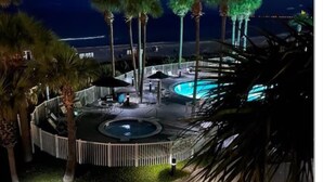 A nighttime view of a circular pool area with palm trees and lounge chairs, surrounded by a white fence. - Grand Panama 809 Beachfront Escape, Beach Service + Pets OK! (Panama City Beach)