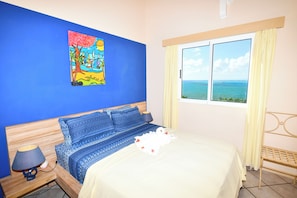 Une chambre à coucher avec un lit, une table de chevet, une lampe, une fenêtre avec des rideaux et un tableau au mur.