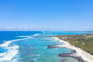 Eine Luftaufnahme einer Küstenregion mit klarem, blauem Wasser, einem Sandstrand und einem bewaldeten Gebiet mit Gebäuden.