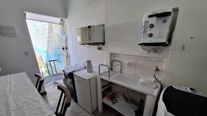 Una cocina compacta con un lavamanos, microondas y un refrigerador pequeño. Un comedor con sillas está ubicado cerca de la ventana, la cual ofrece vistas al exterior.