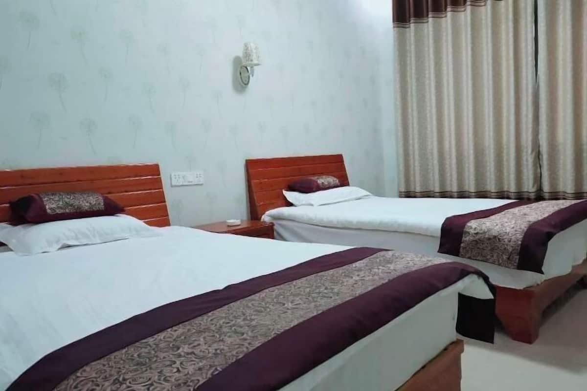 Une chambre d’hôtel avec deux lits, chacun doté d’une tête de lit en bois et de linge de lit blanc. Une table de chevet avec une lampe se trouve entre les deux lits. Les murs de la chambre sont de couleur claire et présentent un motif floral. Il y a également une fenêtre avec des rideaux.