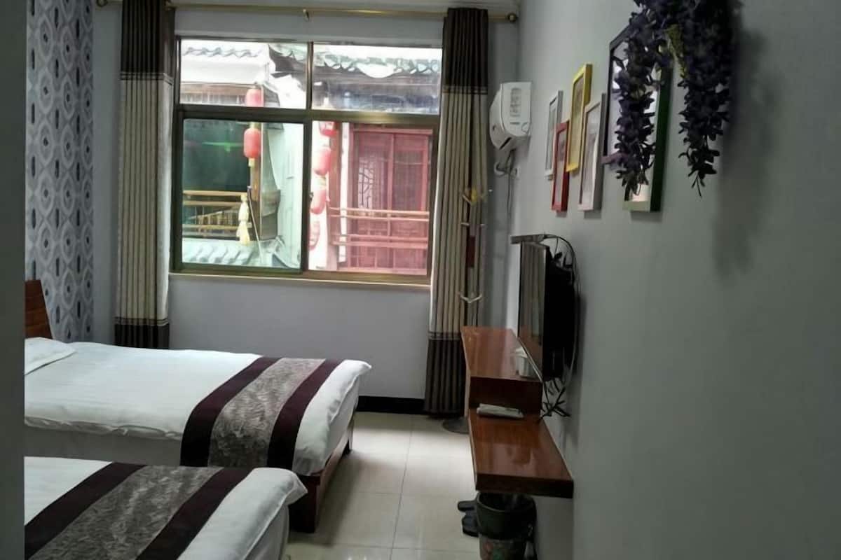 Une chambre d’hôtel avec deux lits, un bureau en bois, une fenêtre donnant sur un bâtiment traditionnel et un climatiseur fixé au mur.