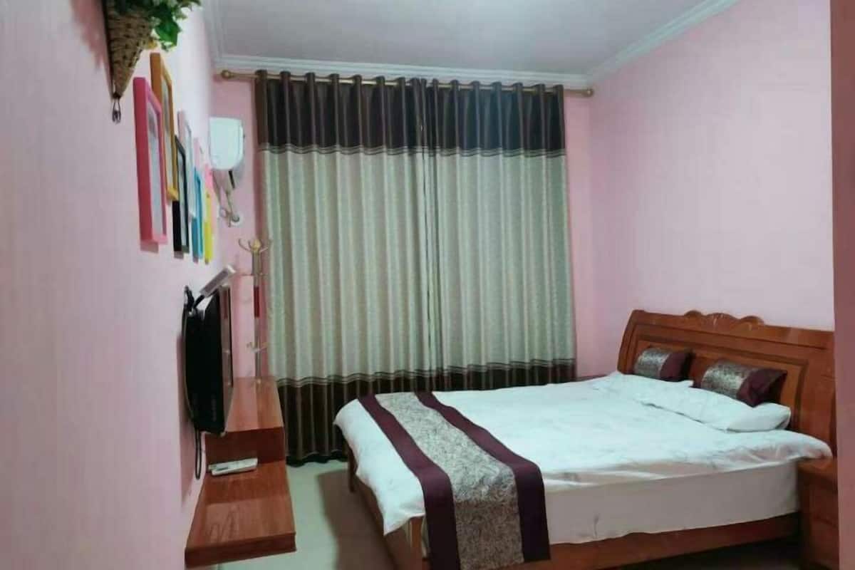 Une chambre à coucher avec un lit, une tête de lit en bois, une table de chevet, une plante suspendue et un rideau.