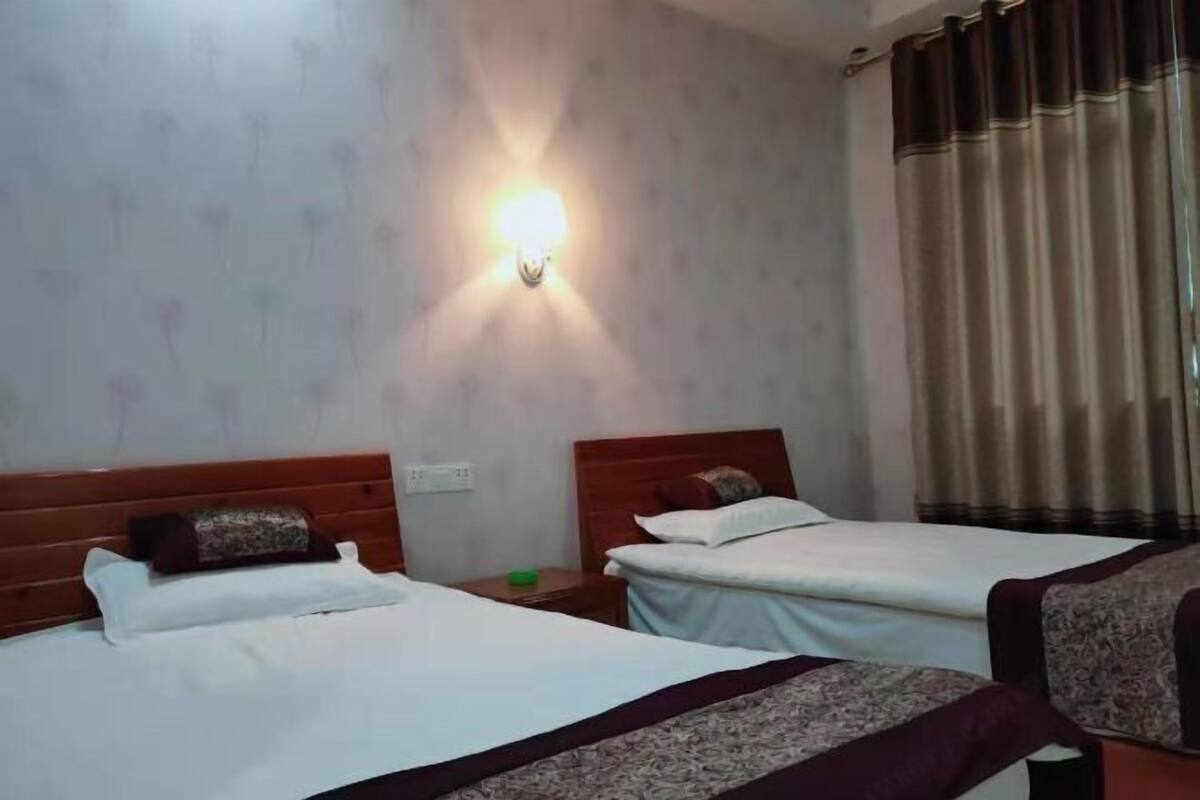 Une chambre d’hôtel avec deux lits, une lampe fixée au mur, un rideau et une table de chevet sur laquelle se trouve un objet vert.